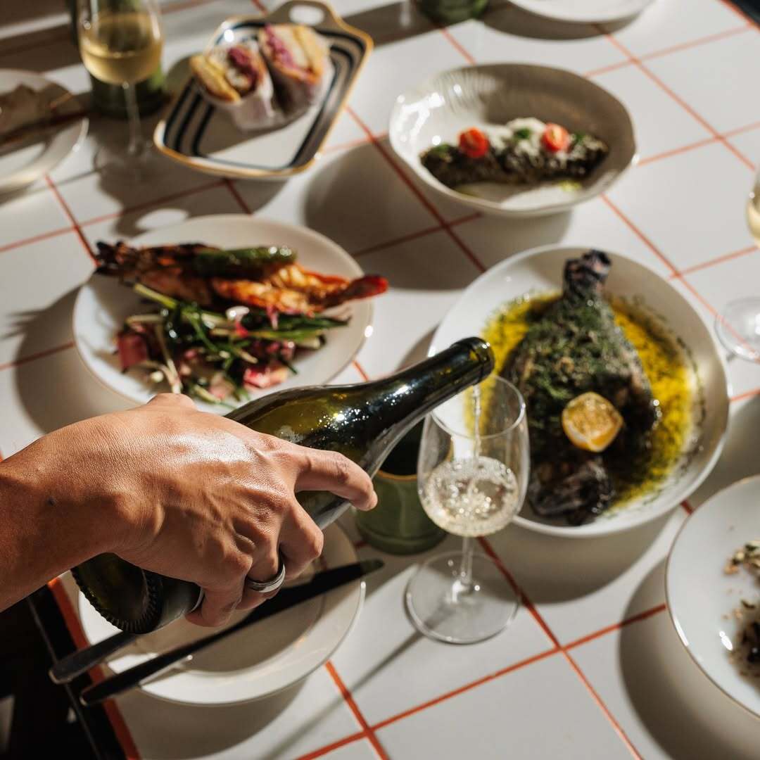 Wine poured at a table of Bar Abong dishes, including grilled prawns and whole fish in herb oil